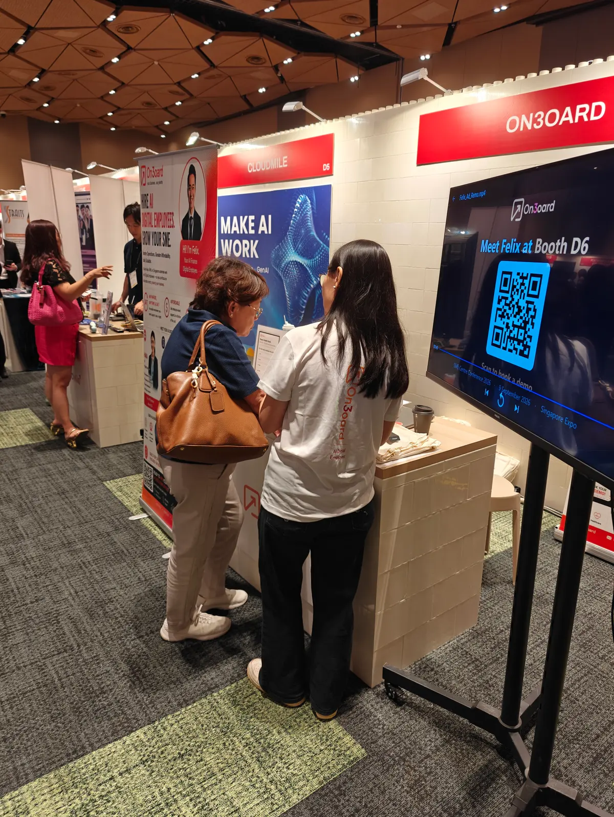 A visitor and an On3oard team member at the booth counter, with a screen reading Meet Felix at Booth D6 — scan to book a demo
